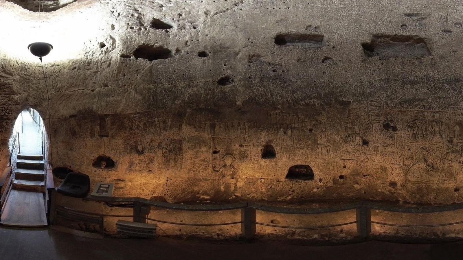 A lit up view of the cave, showing the museum entrance on the left side with a brighter corridor and four steps down into the cave. The walls are white and chalky, lit by an uplighter. Many things are etched into the wall, which has several pockets dug out, and there are wooden/rope barriers in front of the carvings.