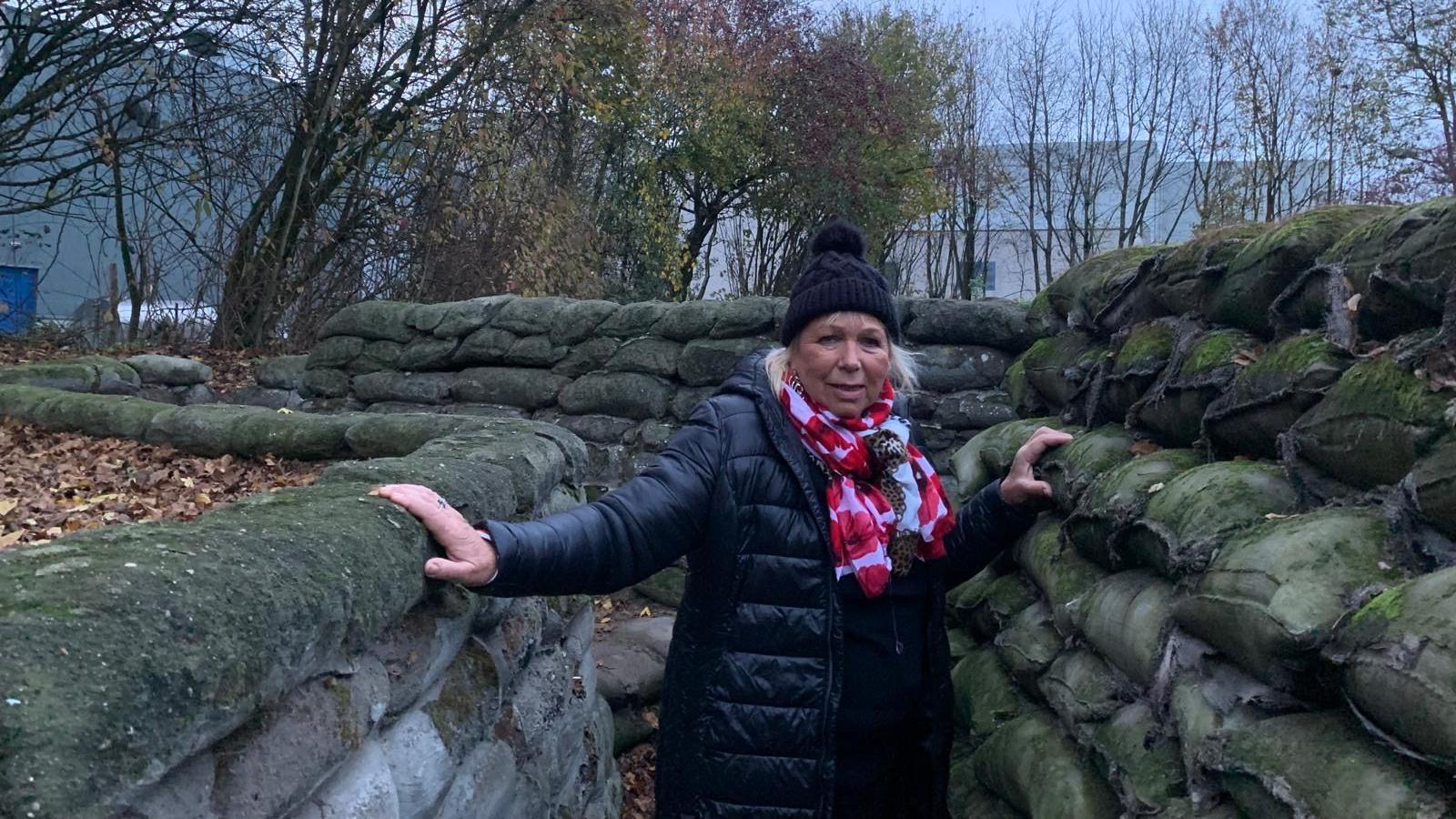 Sadie Nine, standing in a World War One trench, wearing a black coat, black hat, a scarf with her two hands resting on the top row of the sides of the trench. Sandbags are all around her and trees are in the distance.