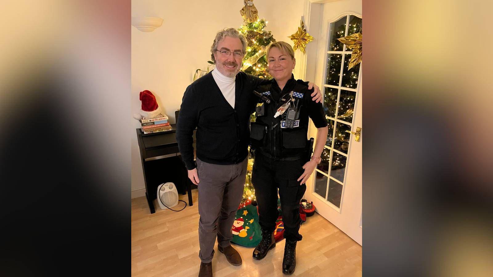 Peter Prendiville is smiling and has his arm around PC Francolini shoulder. Mr Prendiville has white curly hair, wearing thin framed glasses, a white turtle neck under a black cardigan with grey trousers and brown suede Chelsea boots. PC Francolini has blonde hair in a bun and is wearing all black police uniform.