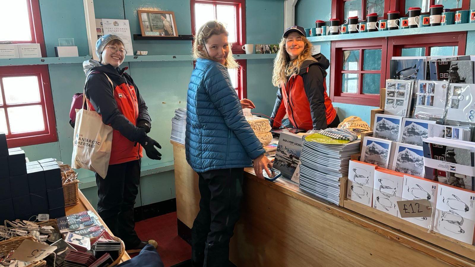 Three people - Aoife, Lou and Dale - in a gift shop. They are surrounded by cads, mugs and other souvenirs 