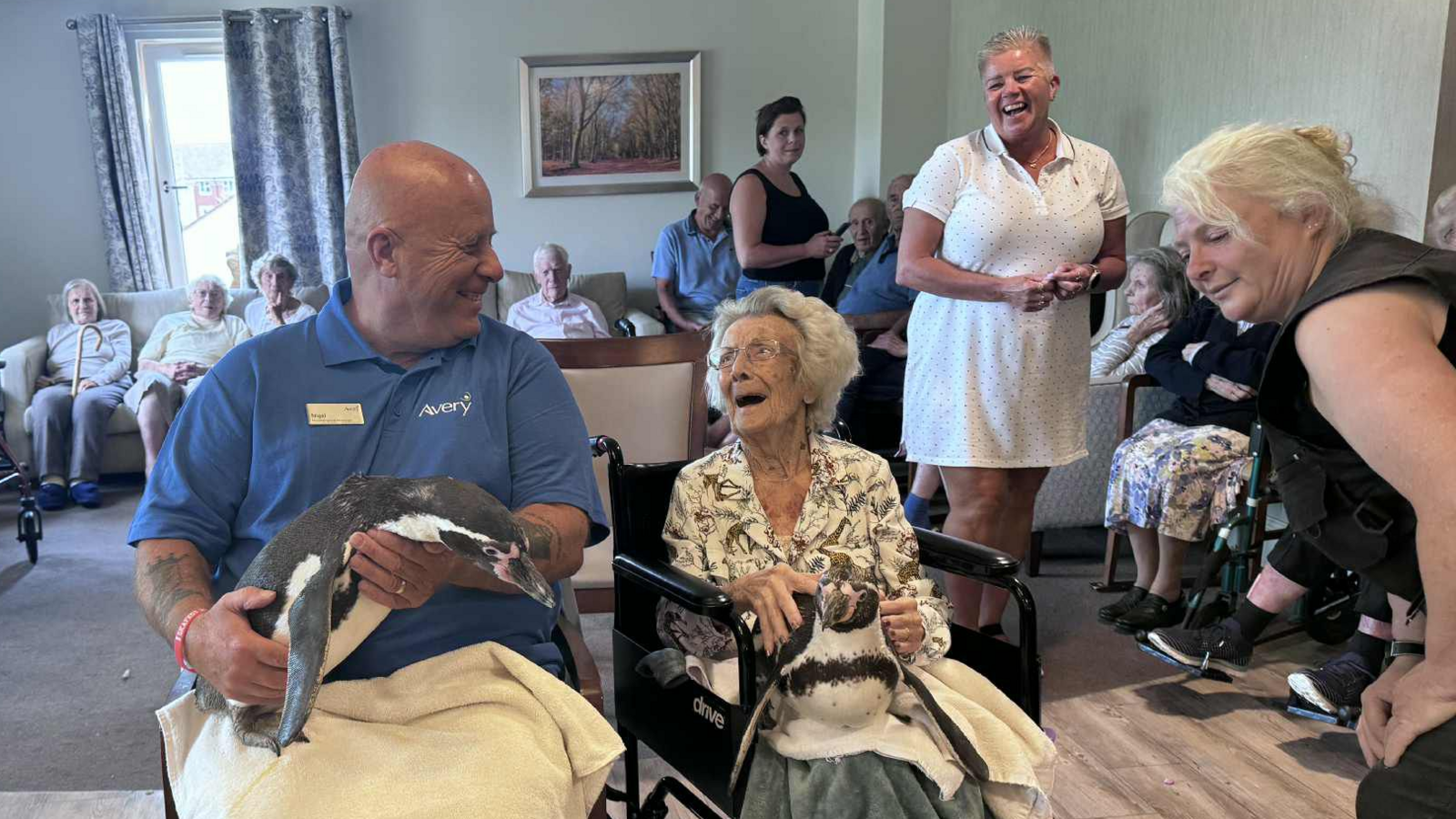 An elderly woman laughs as she sits with a penguin on her lap. Next to her is a man in a blue polo shirt who is also holding a penguin. In the background are other care home residents and staff.