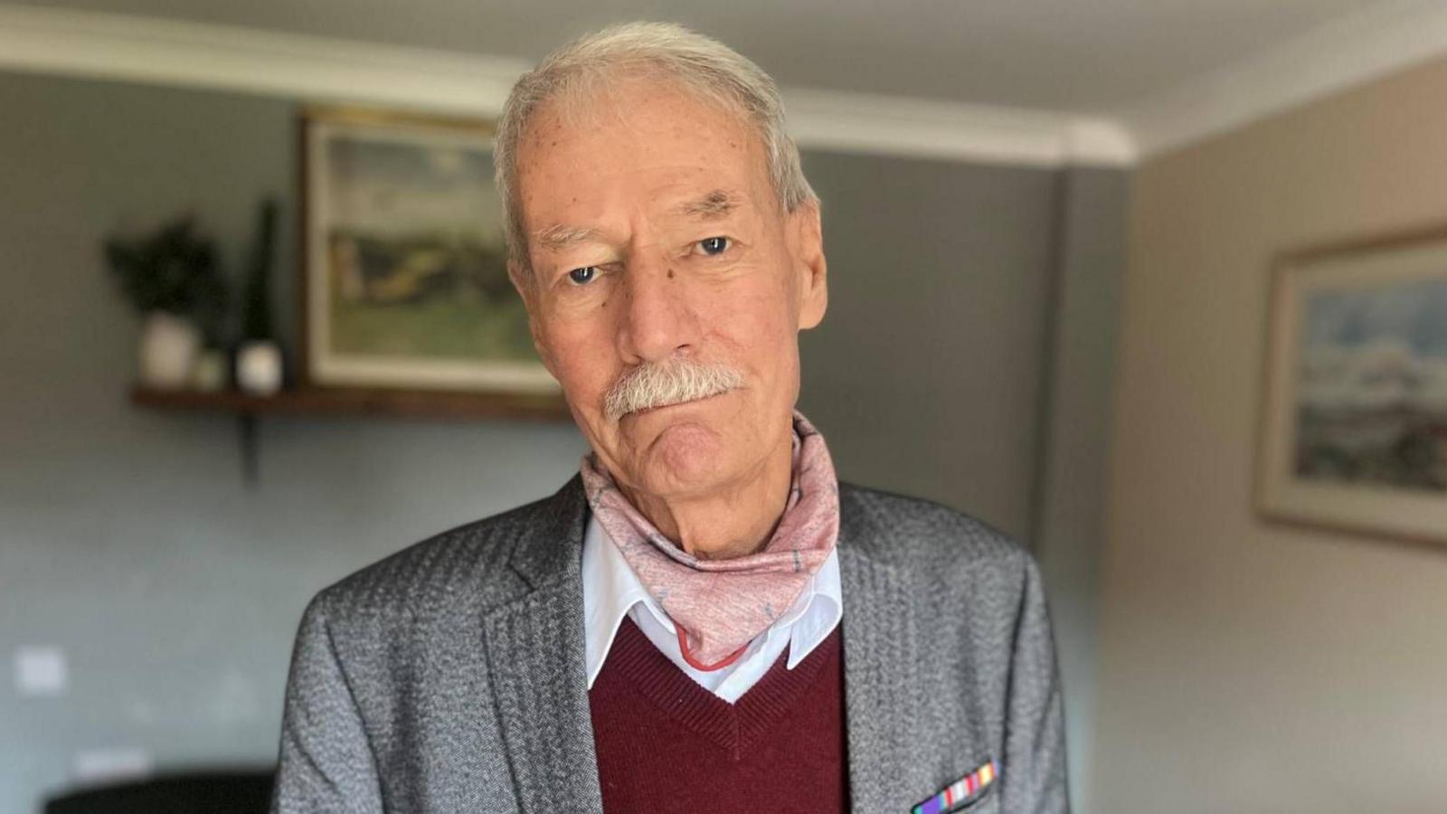 Robert James has light grey hair and a moustache. He stands in a room wearing a grey jacket, Burgundy sweater, white shirt and pink scarf.