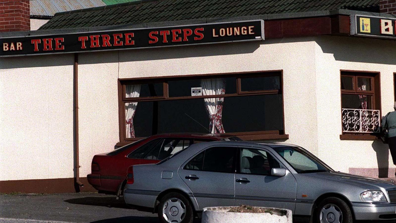 An archive photo of the Three Steps pub in Dromintee, County Armagh.   The building is painted cream with brown windows, with pub signs on the facia boards.  Two cars are parked outside.