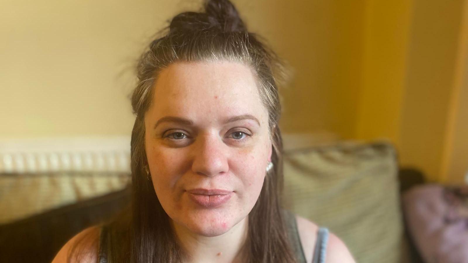 A woman with brown hair looks straight at the camera while sitting on a sofa.