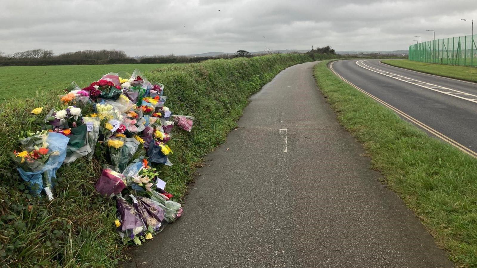 Tens of bunches of colourful fresh flowers paying tribute to Samuel Vass are paid on a hedge on a path next to a road. Some of the high fencing surrounding RNAS Culdrose can be seen to the right of the road.