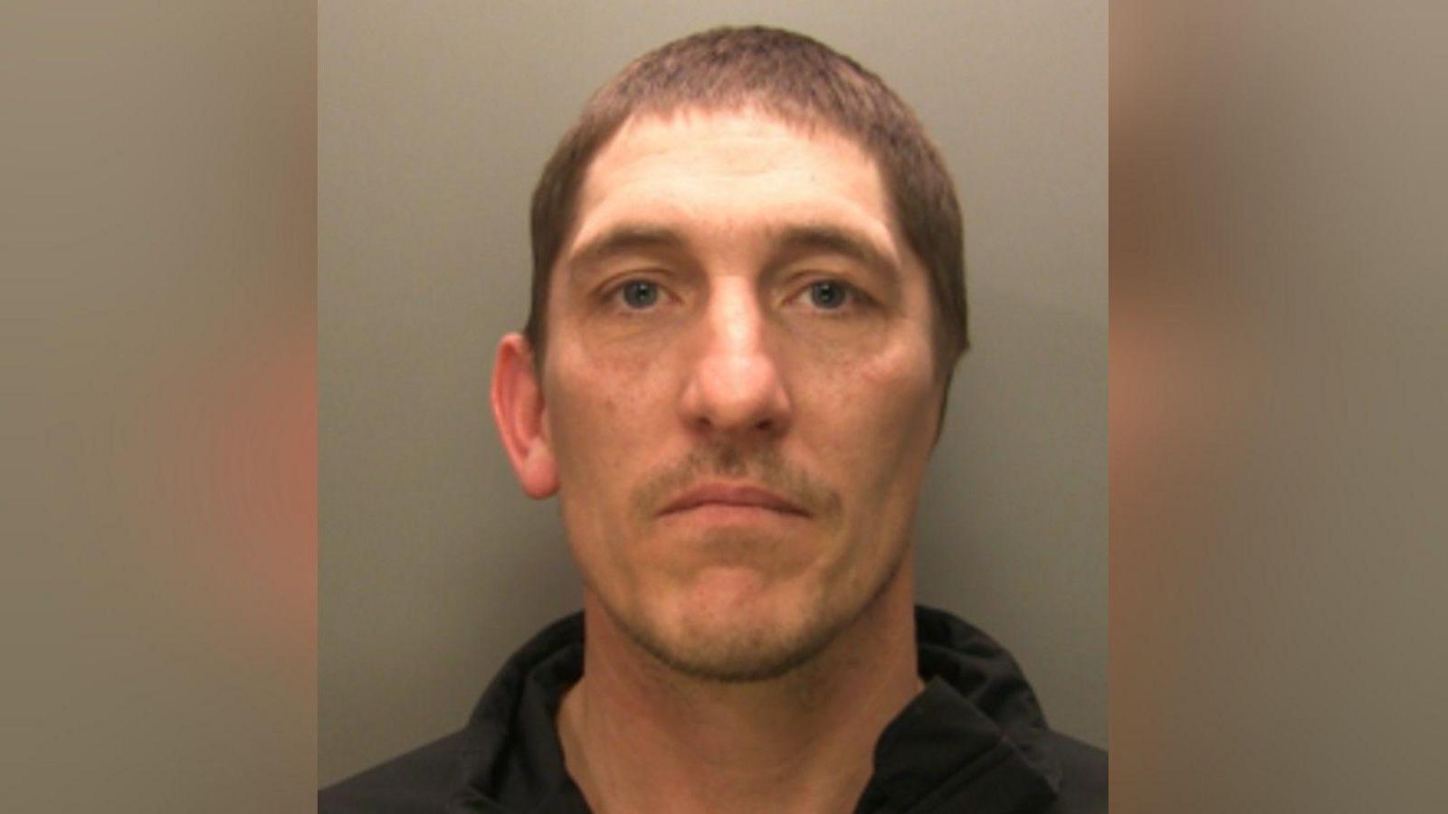 A mugshot of Christopher Hewer. He has short brown hair and blue eyes and his wearing a black hoodie. He only has one ear.