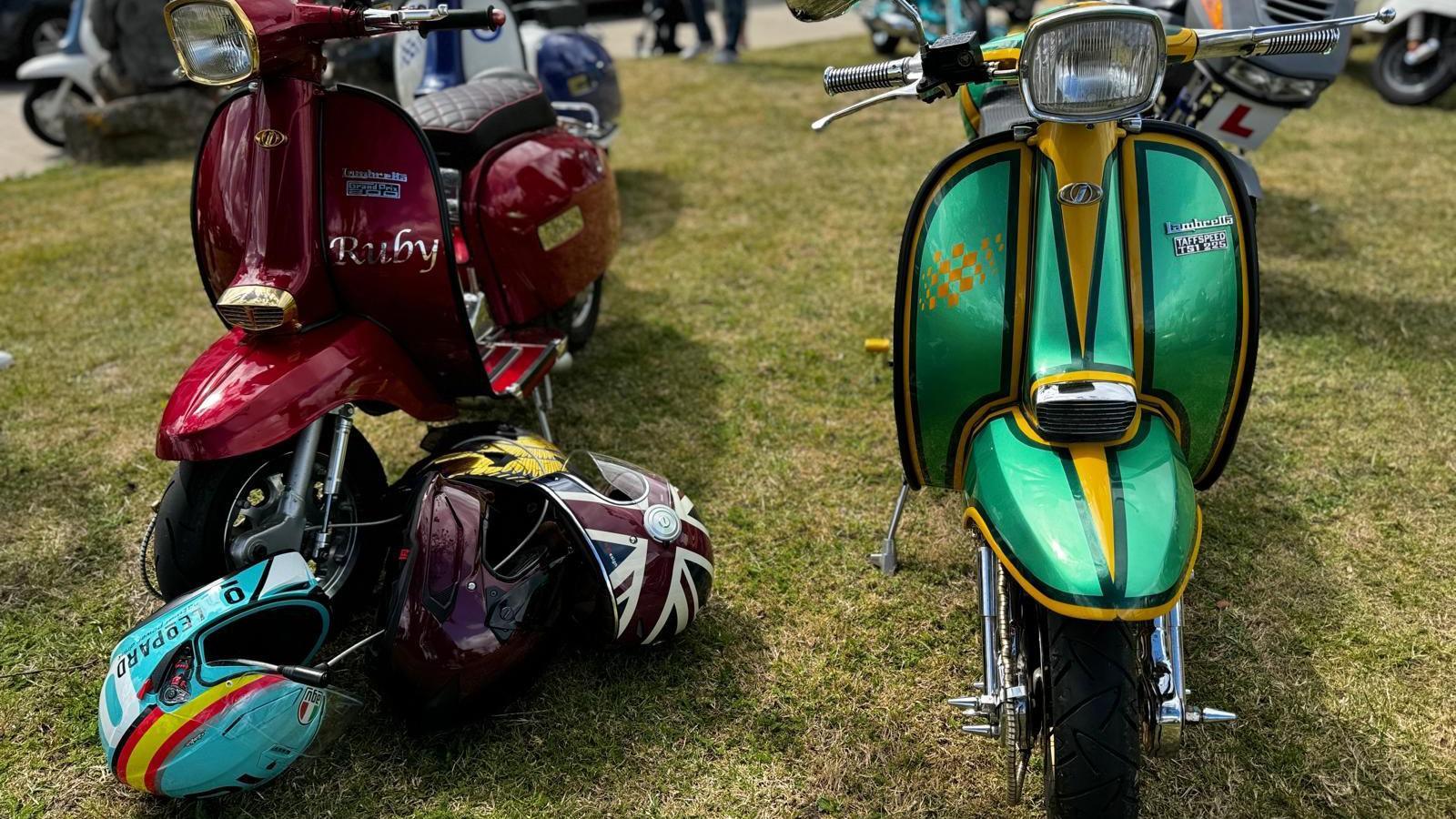 Two customised Lambretta scooters. One is metallic deep red with the word Ruby in metallic lettering on the faring. The other is metallic green with a dark green and gold detailing around the edge