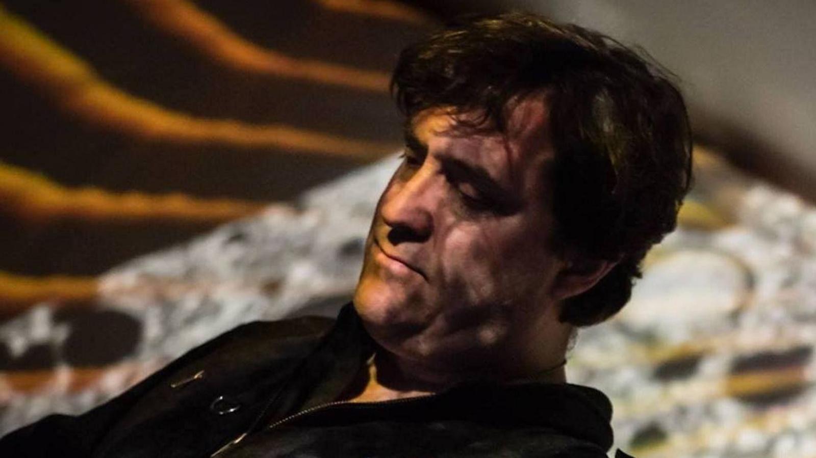 Richard Norris is looking down to left of the picture while DJ'ing at a music venue. He has unkempt black hair and there is colourful lighting across his face and on the backdrop.