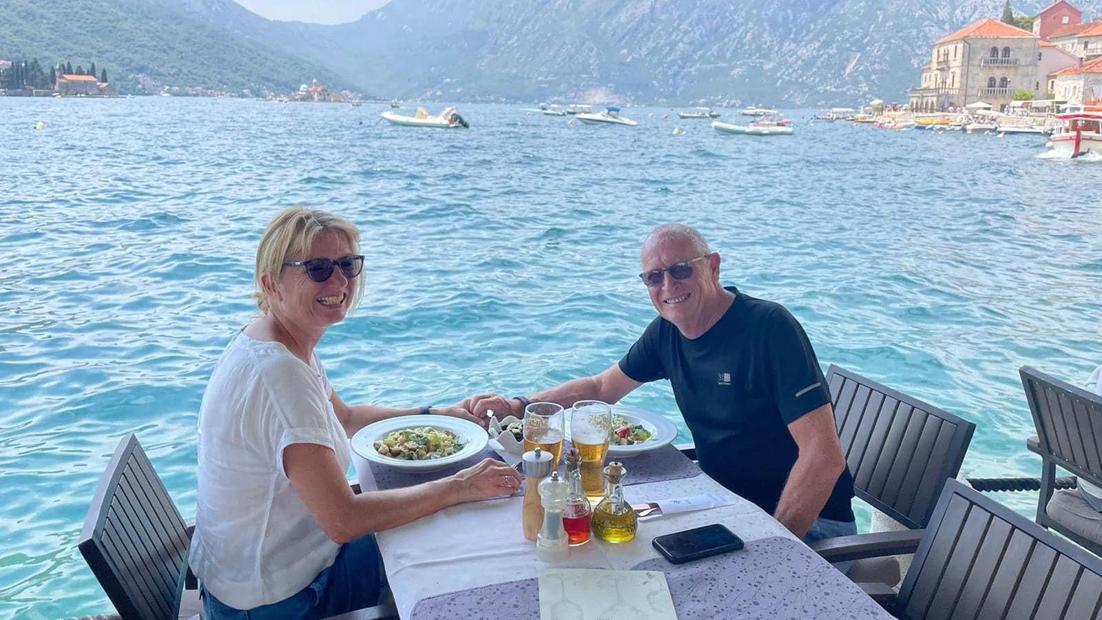 Jill and husband at a table in front of a wide blue lake, with villas on the distant shore and mountains behind the water. There are boats and dinghies dotted across the water.