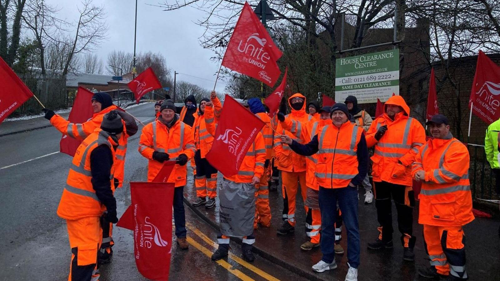 A group of men wearing high-vis orange jackets and trousers stand on a picket line waving red Unite the union flags, next to a road near the canal.