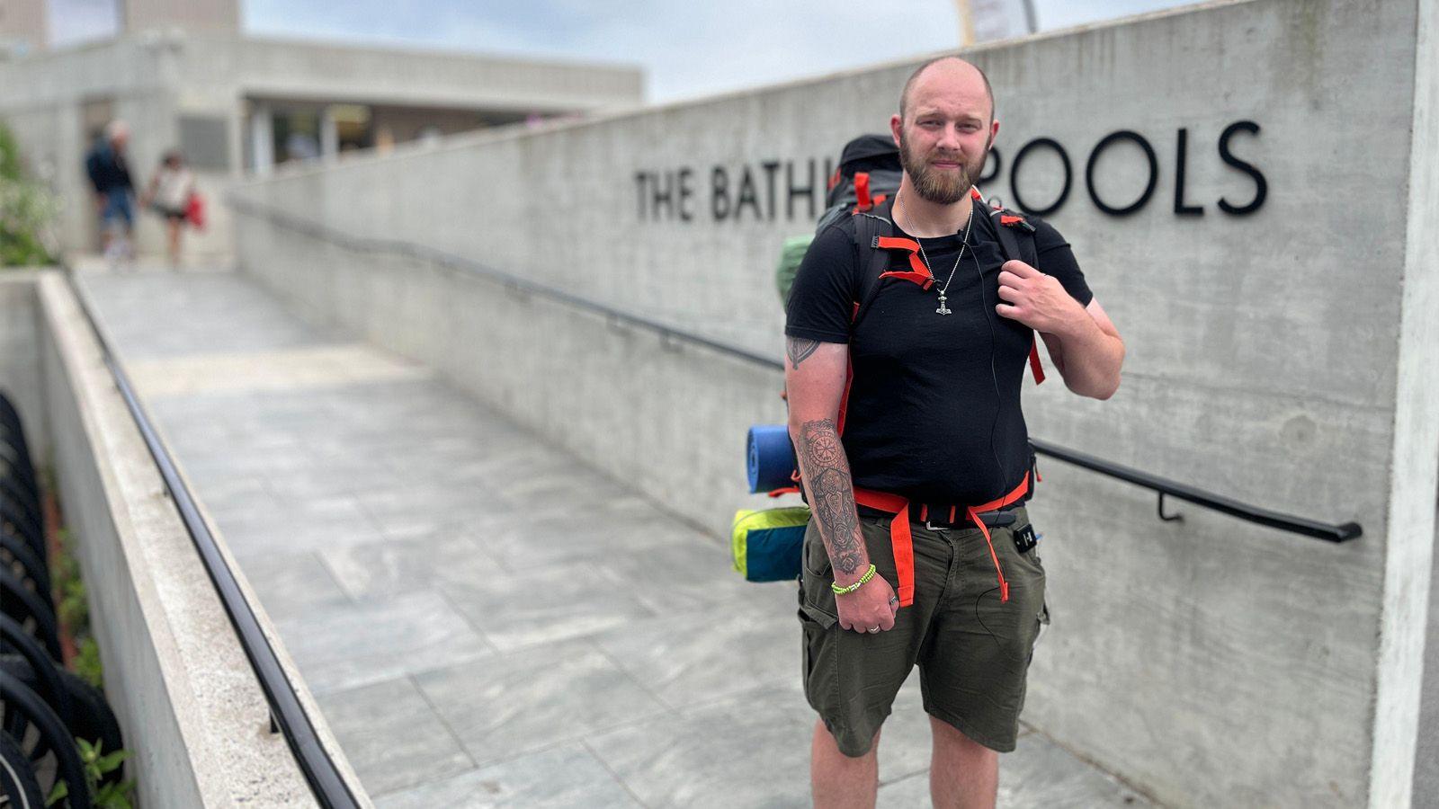 A bald, bearded man in his late 20s stands in front of a brutalist building and a The Bathing Pools sign. He has tattoos on his forearm, wears a black T-shirt and green shorts, and carries a large backpack.