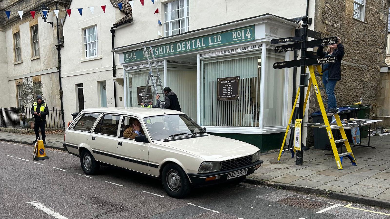 An old estate car is parked in front of traditional-looking white painted cotswold stone building, which has a shop front. The sign on it reads Rutshire Dental. A member of the crew is on a ladder to the side, disguising a signpost