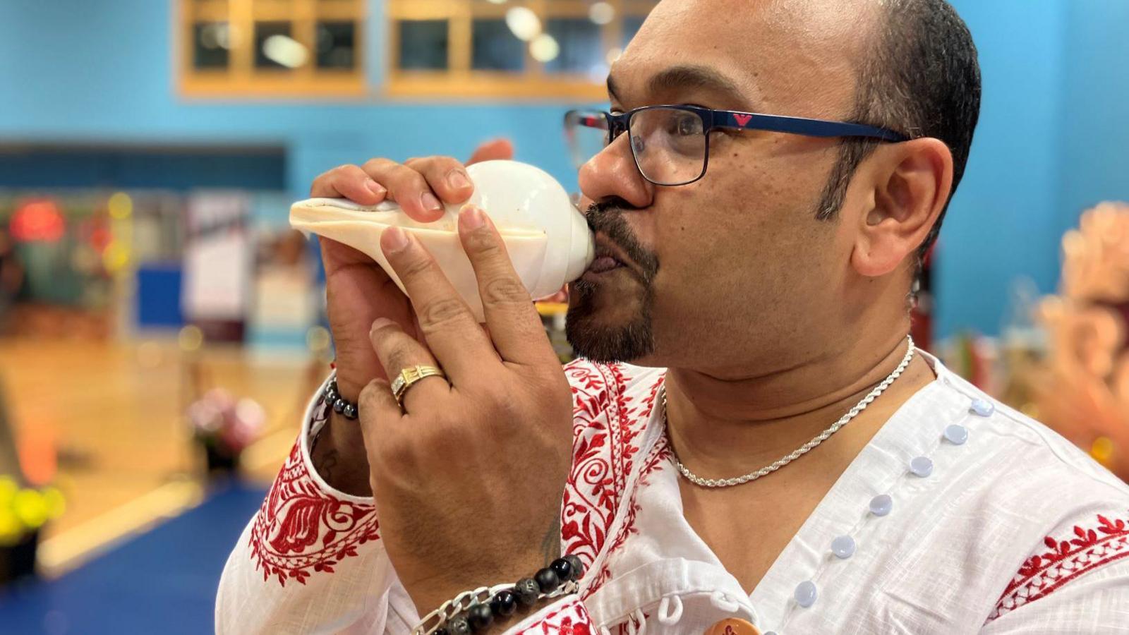 A man blows into a white conch shell. He is wearing traditional Bengali clothes