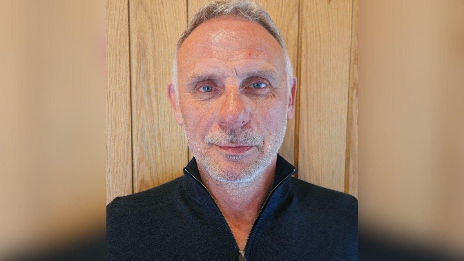 Shaun Adams is pictured with a neutral expression against a wooden door. He has balding grey hair and a white stubble beard. He is wearing an open zip-up navy jumper.