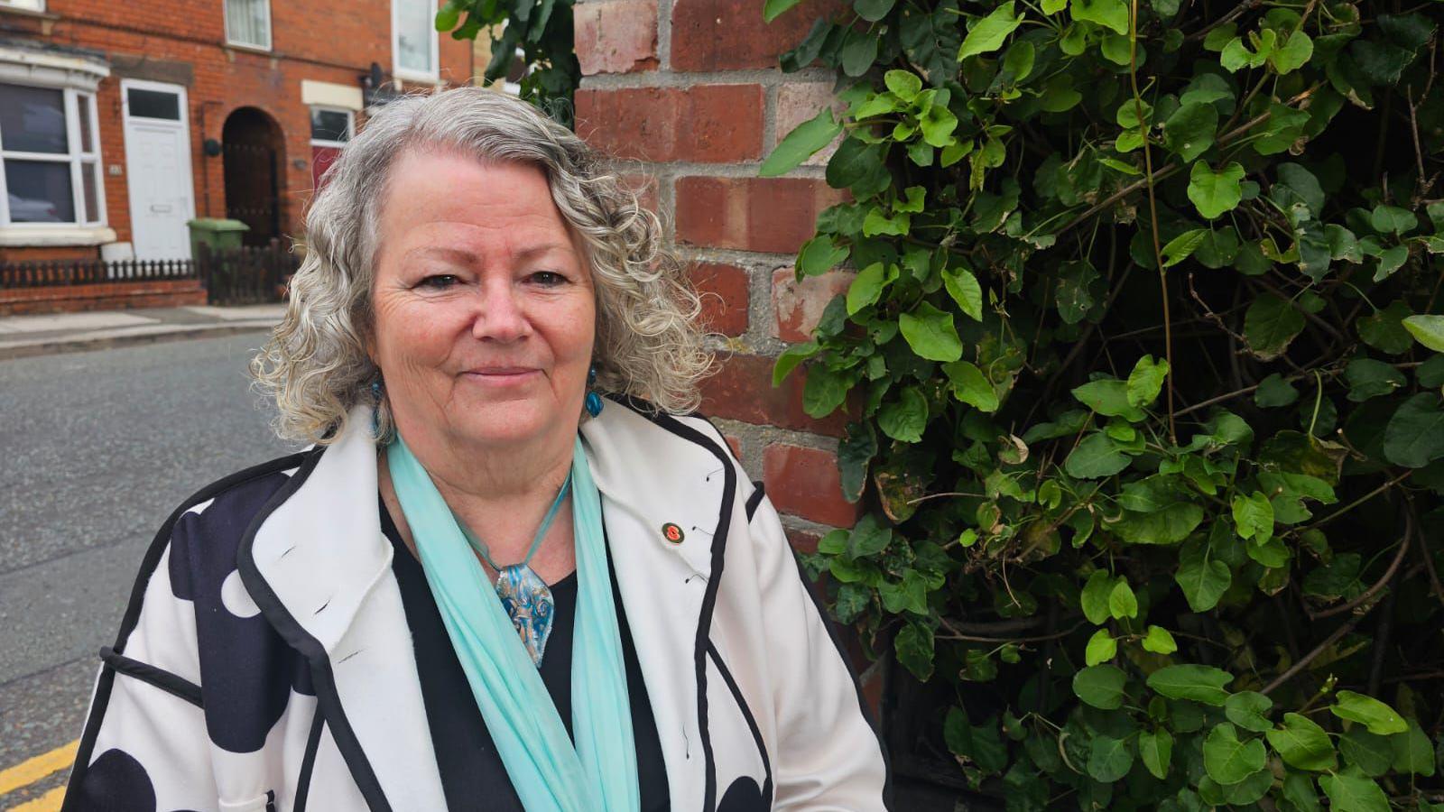 Diane Spurr has grey white curly hair, she is wearing a blue scarf and black and white printed jacket and is smiling at the camera. 