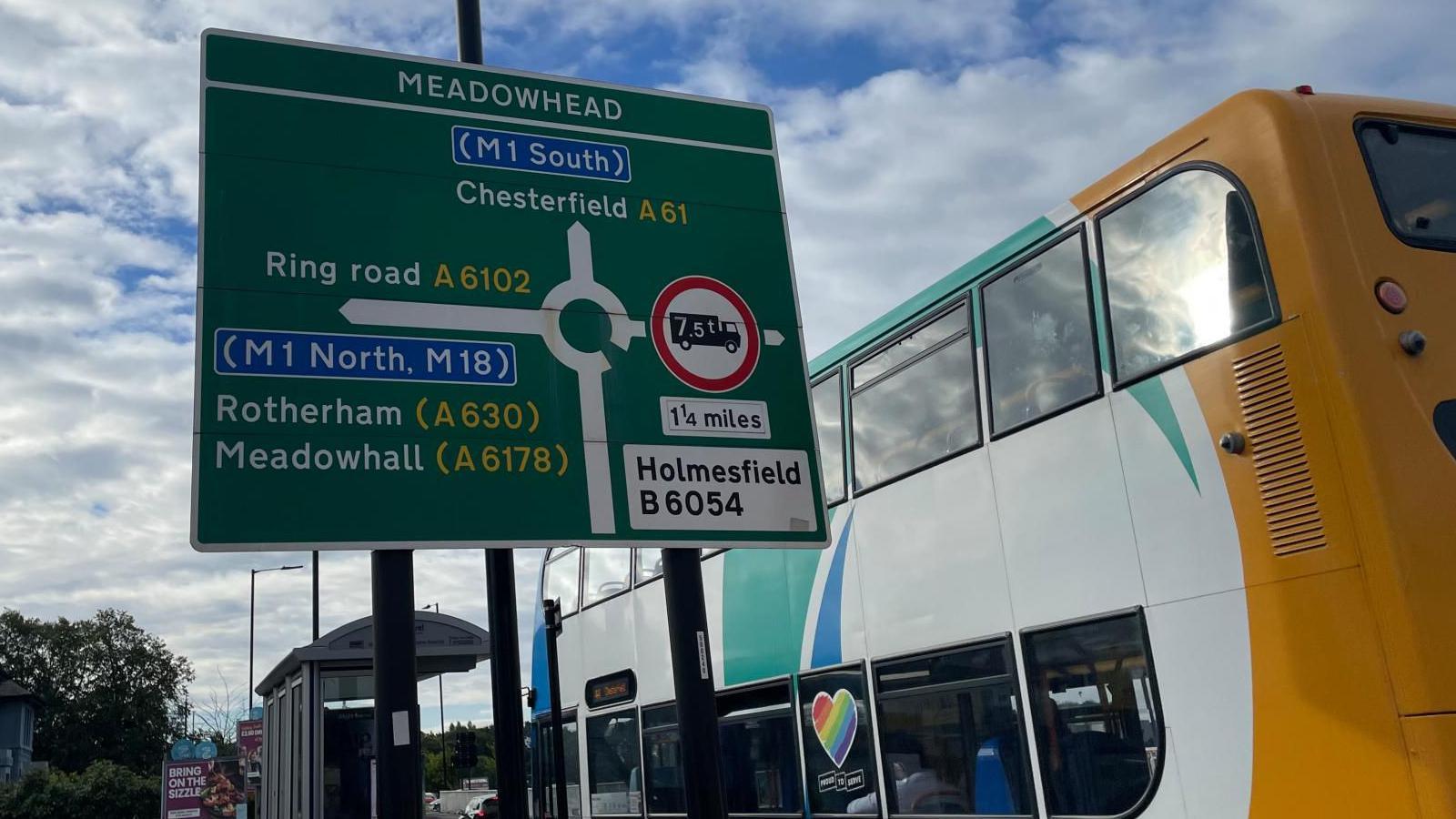 A large road sign showing a roundabout and a bus pulling up to a bus stop.