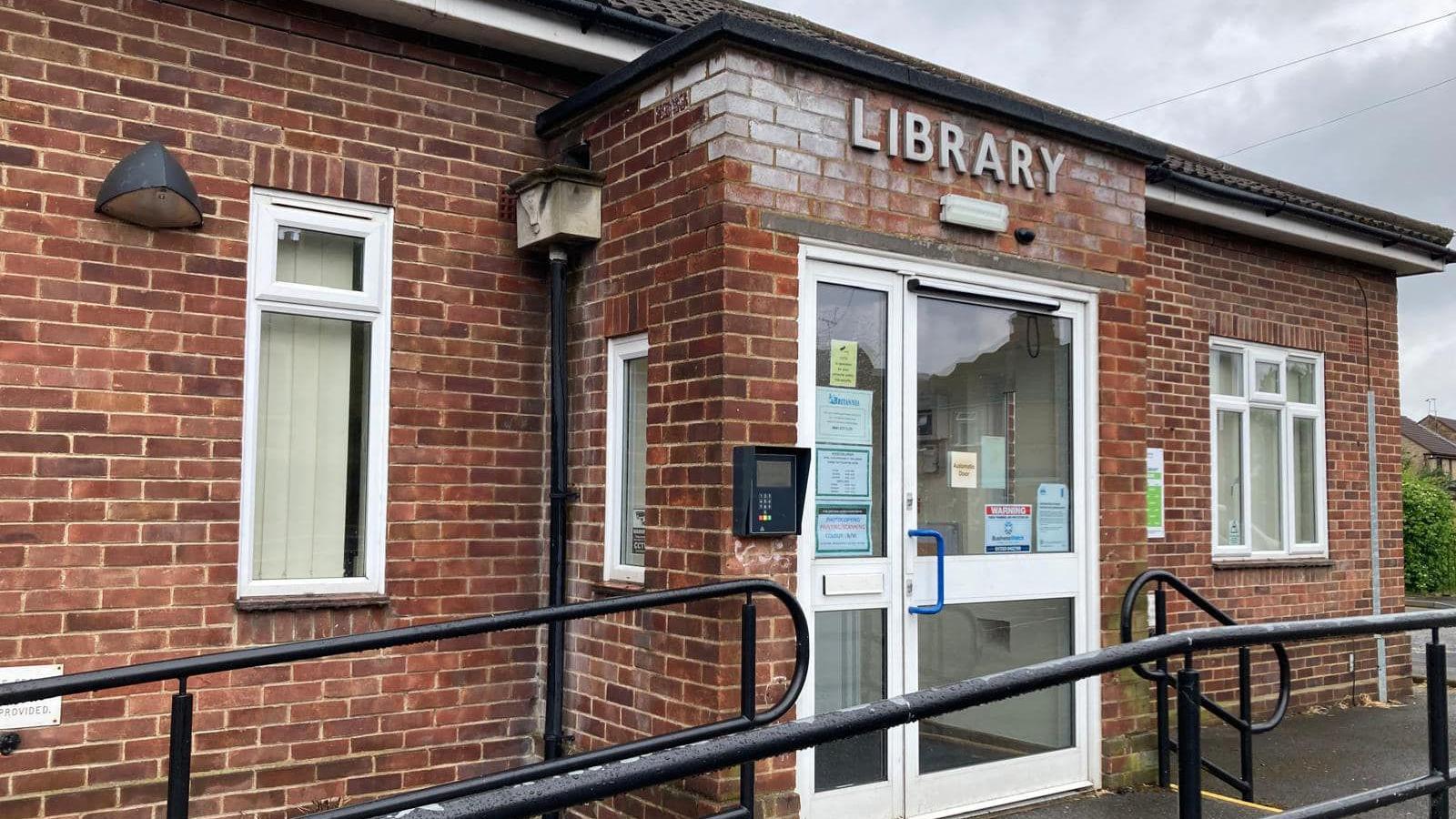 One-storey red-bricked building with a silver sign saying library and access railings leading up to the door.