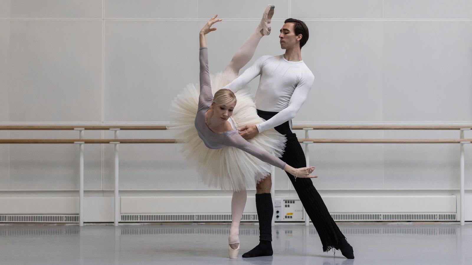 Melissa dancing with Gareth Haw in a dance rehearsal studio. She is wearing a grey leotard, white tutu, white tights and white ballet shoes. Gareth is wearing a white long sleeve top, black trousers and black socks. He has dark hair. Melissa is en pointe on her left leg with right leg in the air, she is bent forward with her arms at her side. Gareth is straight with his left leg pointed to his side. He is supporting Melissa's waist.