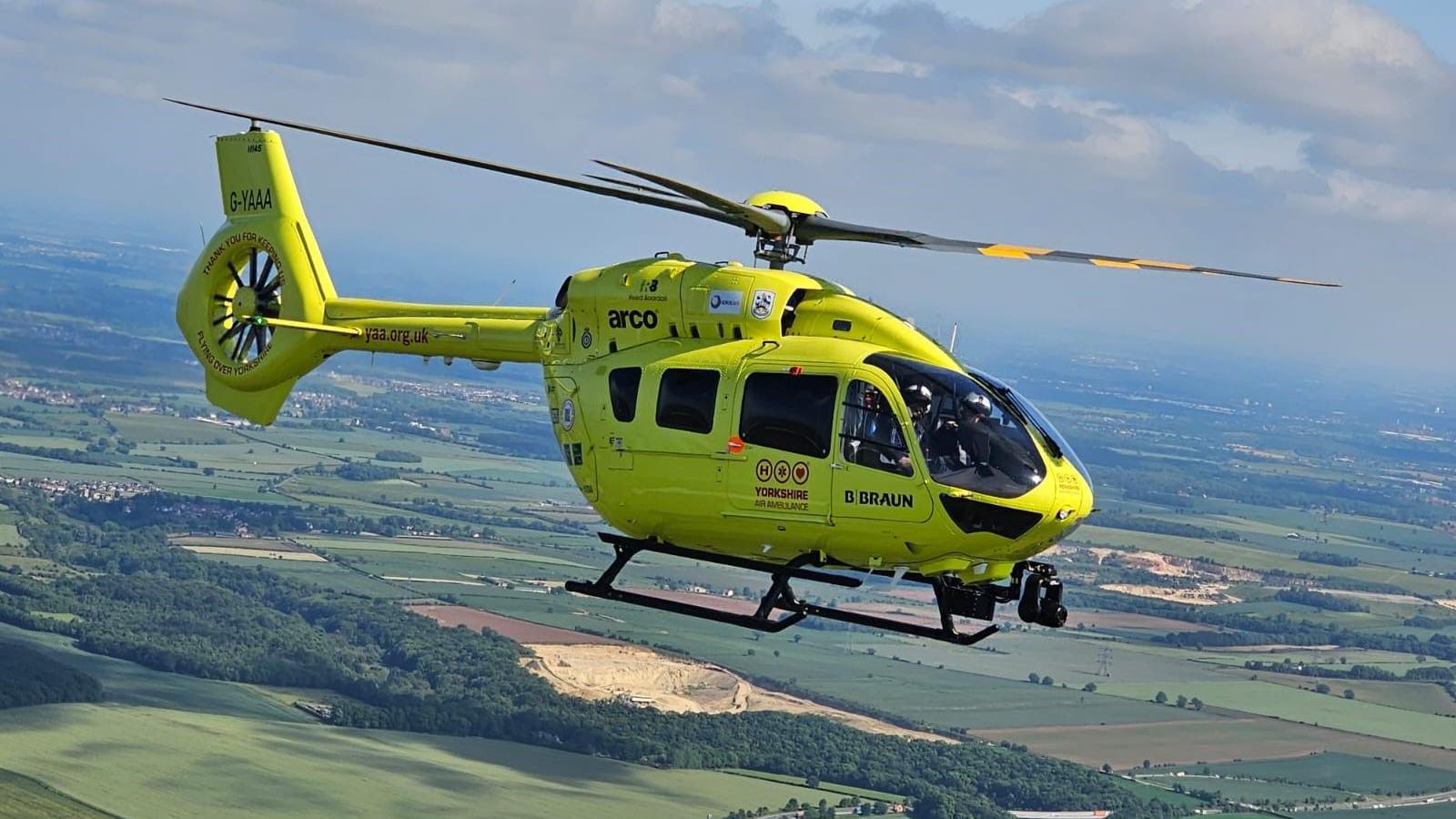 A bright yellow Yorkshire Air Ambulance helicopter in flight. Fields can be seen below. 