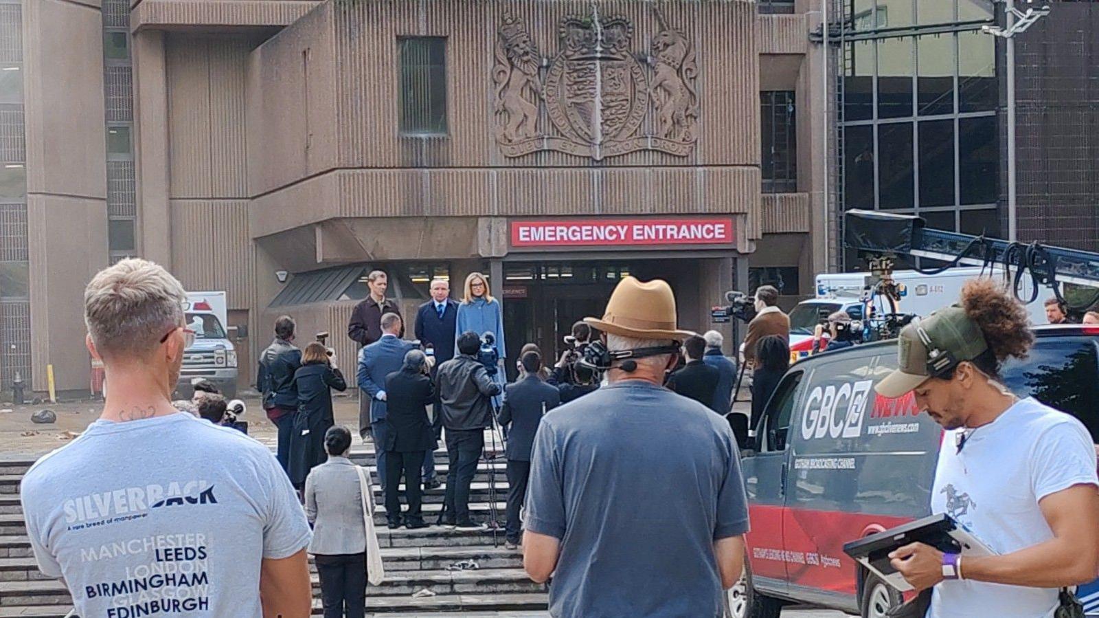 Film crew are stood outside Liverpool Crown Court which is being used as a film set. A red sign across the front of the building says Emergency Entrance in white capital letters. A large amount of film crew are gathered as filming takes place with actors stood on the steps.