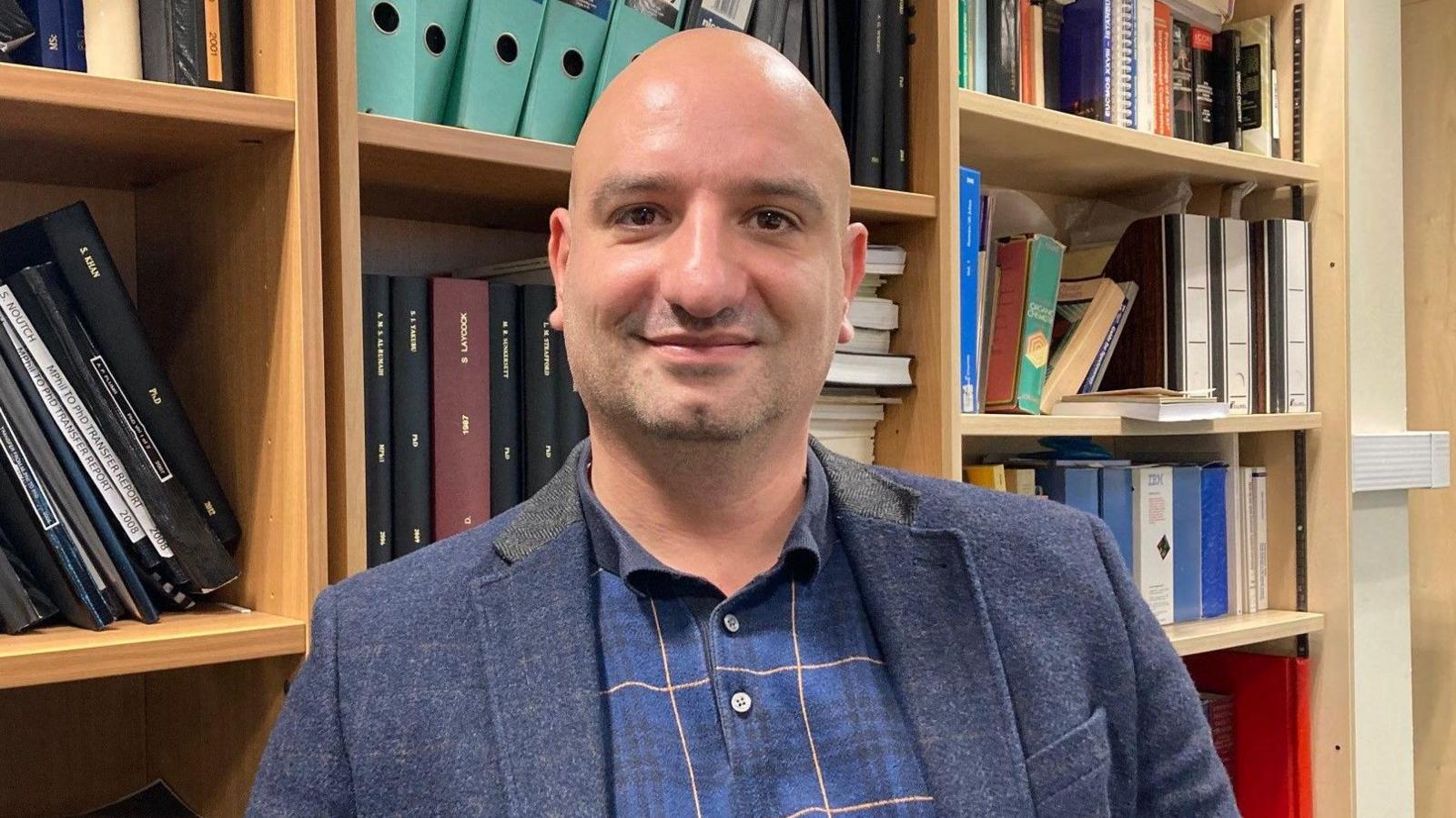 A bald-headed man wearing a blue sports jacket and blue and gold striped shirt sitting down with many multicoloured books on light brown shelves behind him.  