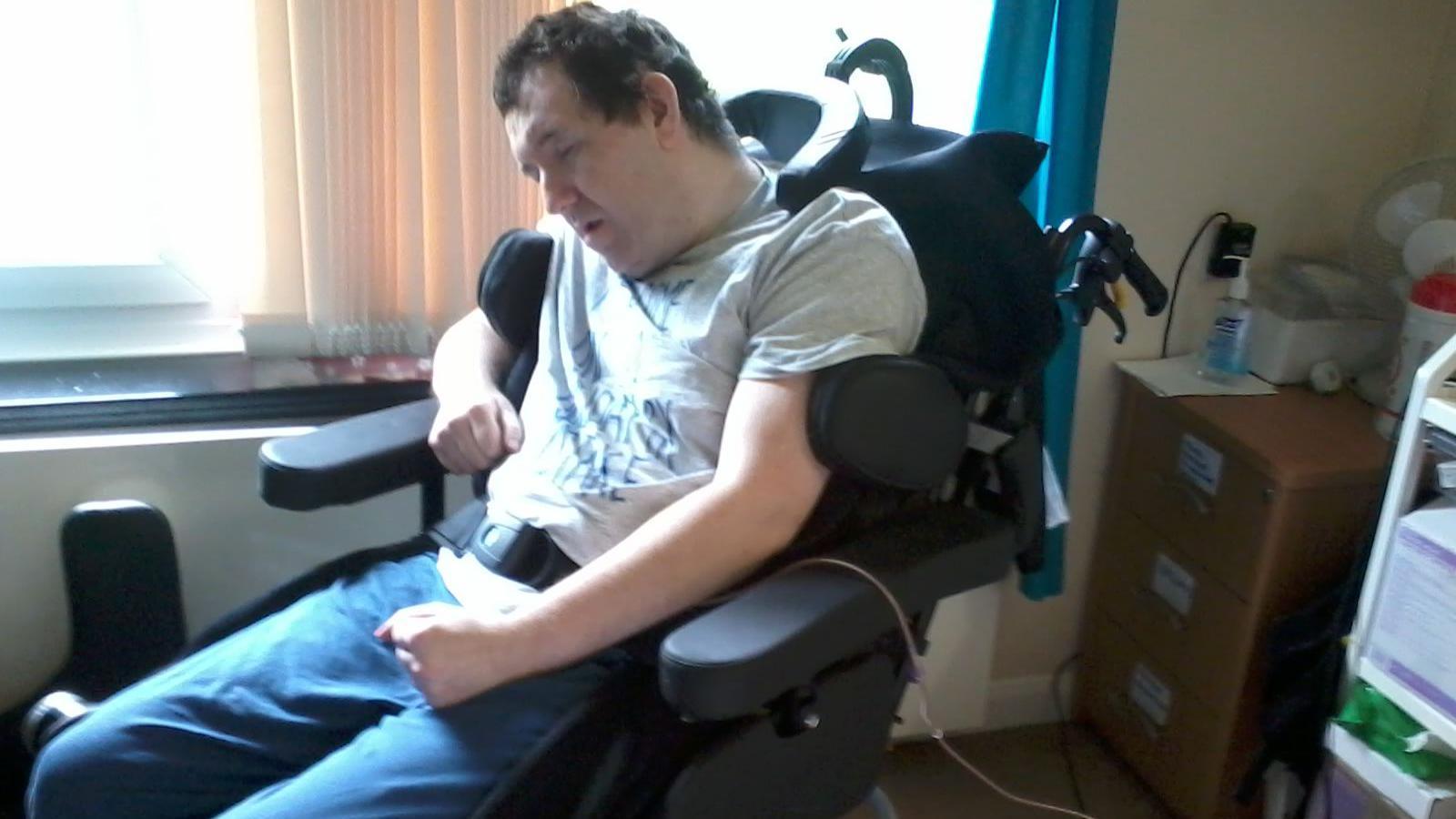 Adam Pickles is in a wheelchair in a house. His eyes are closed and he has a neck support and support for his arms.