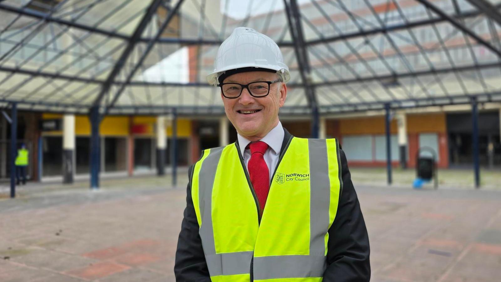 Mike Stonard is wearing a white hard hat and a yellow high-vis jacket with Norwich City Council branding over a black suit and red tie. He is standing under a glass sheltered area on paving.