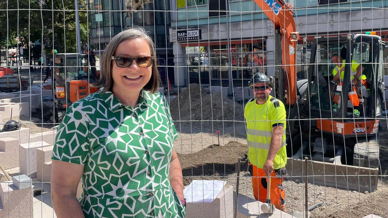 A councillor and a worker, both in shades, smiling either side of a construction site fence