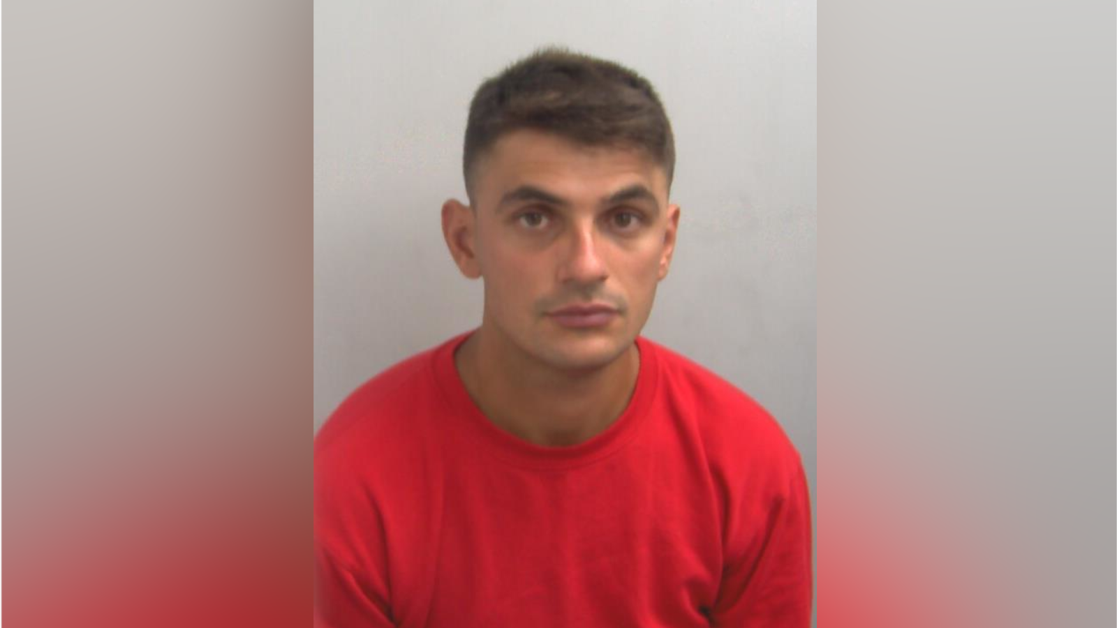 A man wearing a red sweatshirt looks directly into the camera in a police custody picture. He has dark hair and dark eyes. A plain background can be seen behind him.