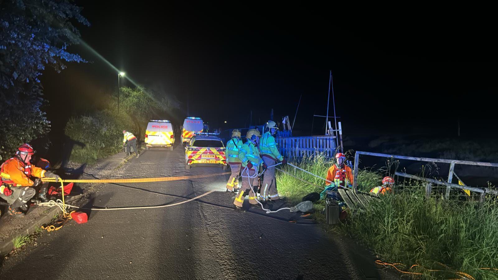 Emergency services teams in hi-vis hackets and safety helmets with specialist equipment to help with the rescue and recovery operation, with three police vehicles seen behind them