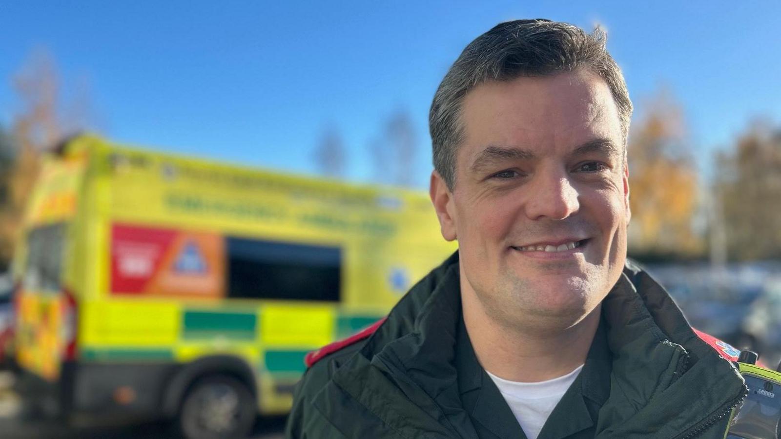 James Atkinson is pictured in his dark green paramedic uniform next to his ambulance. He has short dark hair and is clean shaven. 