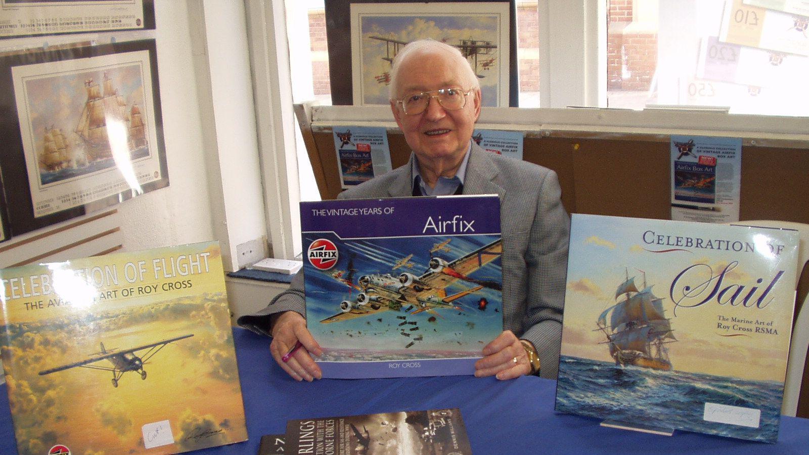 A grey haired man, wearing steel-rimmed glasses and a grey jacket, at a table. He is hold a blue book called "THE VINTAGE YEARS OF Airfix". To the man's right is a yellow book entitled "CELEBRATION OF LIGHT - THE AVIATION ART OF ROY CROSS. To his left is a book called "CELEBRATION OF Sail - The Marine Art of Roy Cross RSMA" 