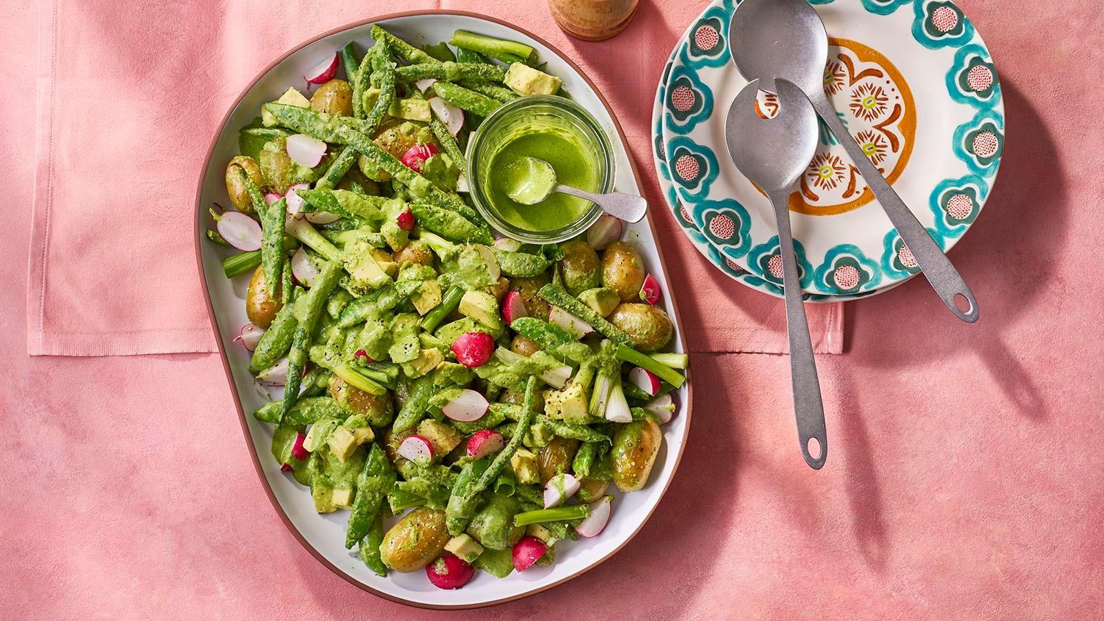 A white oval platter filled with green goddess salad with a pot of green sauce and silver servers on a pink tablecloth