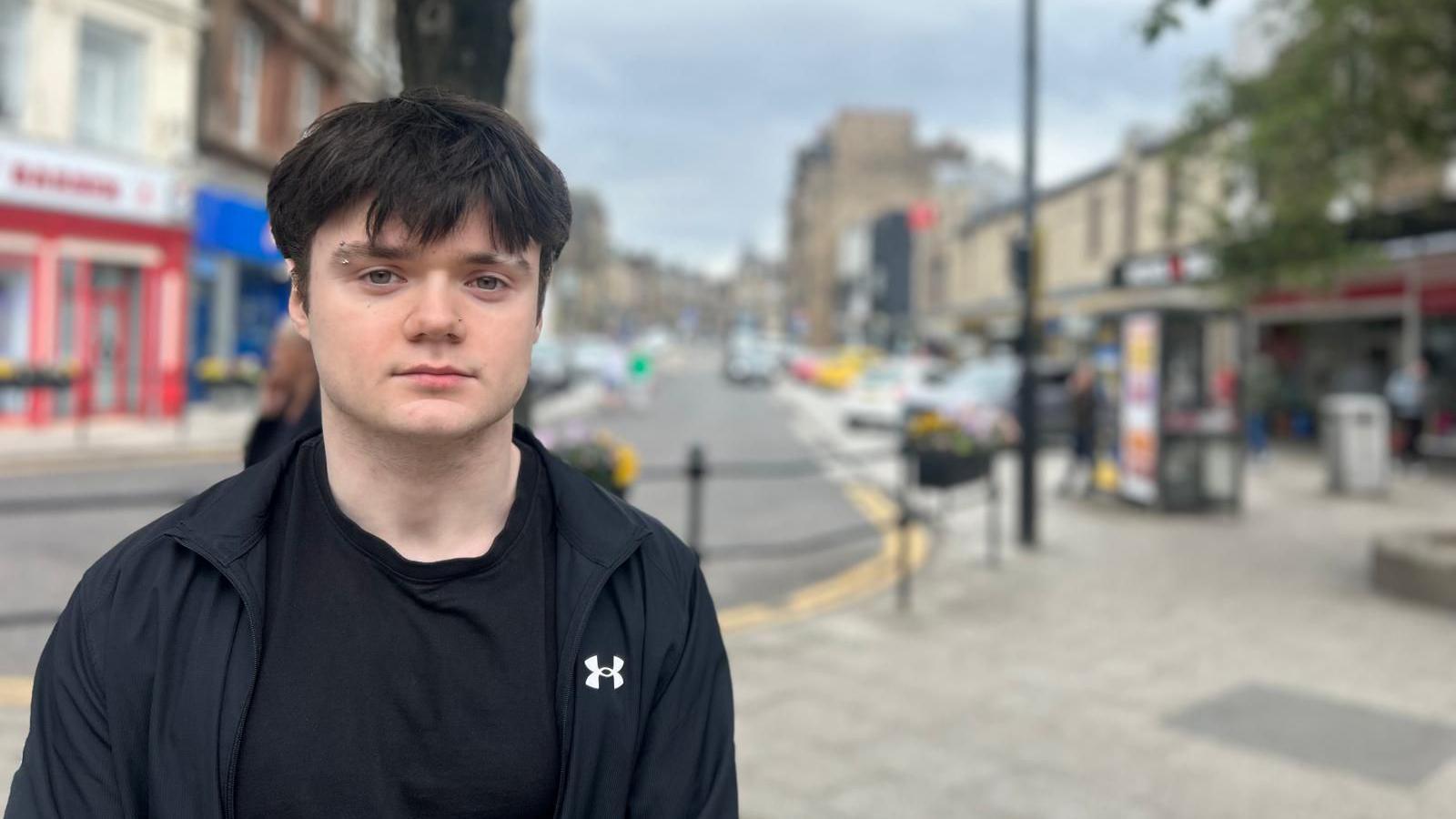 A photo of Alex standing on a high street. He is wearing a black t-shirt and a black tracksuit. He had black hair and a eyebrow stud