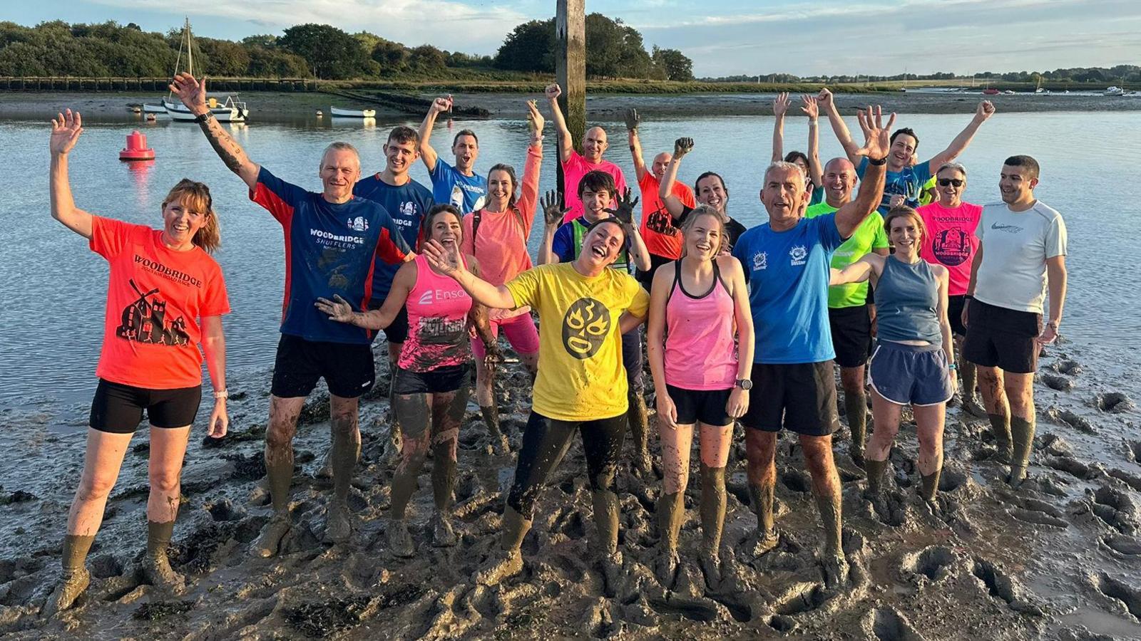 A group of runners are gathered together in the mud and wave and smile at the Camera. They all have muddy legs.