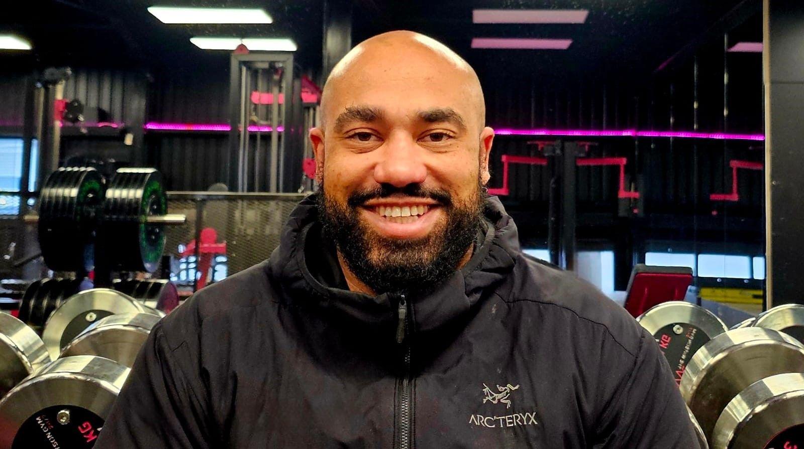 Ram Patten sitting in front of some dumbbells. He is wearing a black coat and is bald with a black beard