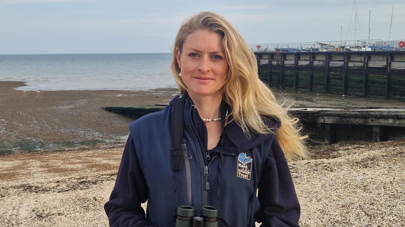 A lady, with flowing long blonde hair; wearing a navy blue fleece; standing on a shingle beach in Whitstable Harbour, with the outgoing tide and windbreaker in the back ground. The lady has a pair of binoculars on a strap around her neck