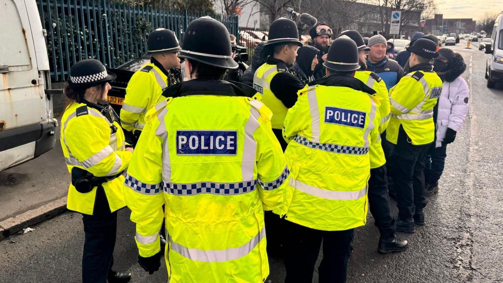 Police officers crowing around protesters on a street. They are wearing hi-vis jackets and there is media in the background. 