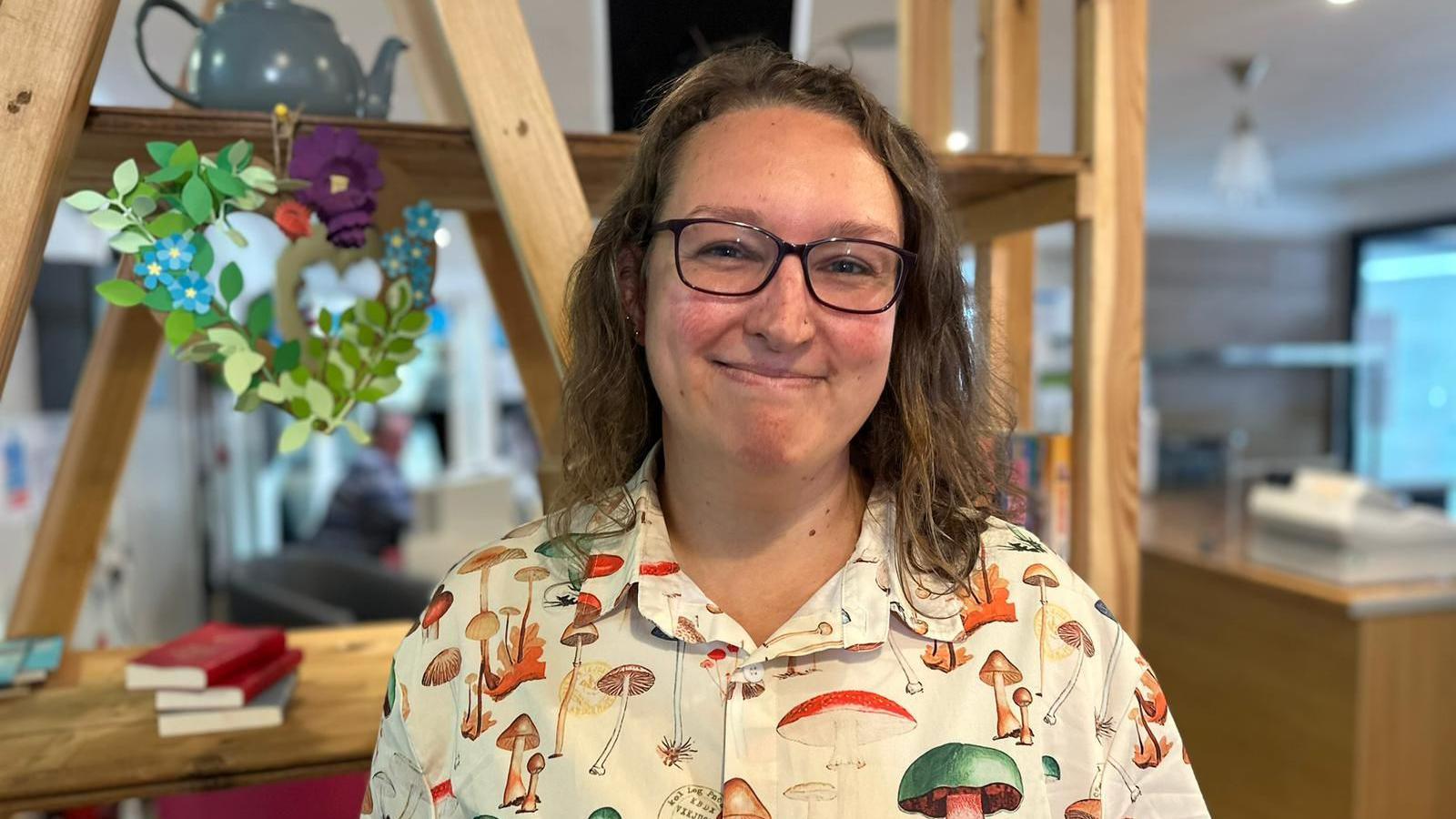 Laura Maggs with shoulder length light brown hair and wearing a cream blouse covered in different sizes, types and colours of mushrooms smiling at the camera with large wooden shelving and office desk in the background
