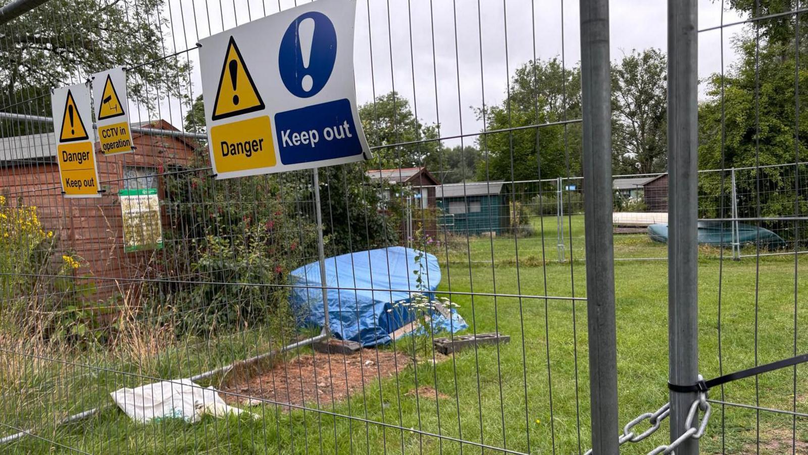 A traditional campsite  with wooden huts is seen  through a metal fence, which has signs saying "Danger" and "Keep Out". Some of the grass is overgrown, and a small boat can be seen covered in a canvas