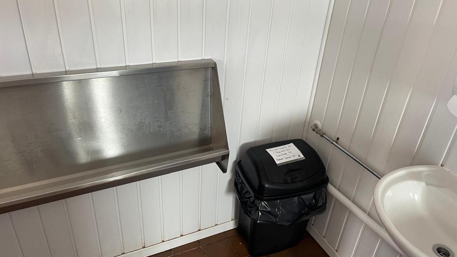 A picture of a small black bin with a piece of paper stuck to it in a corner. The walls are white. There is a sink to the right of the picture.