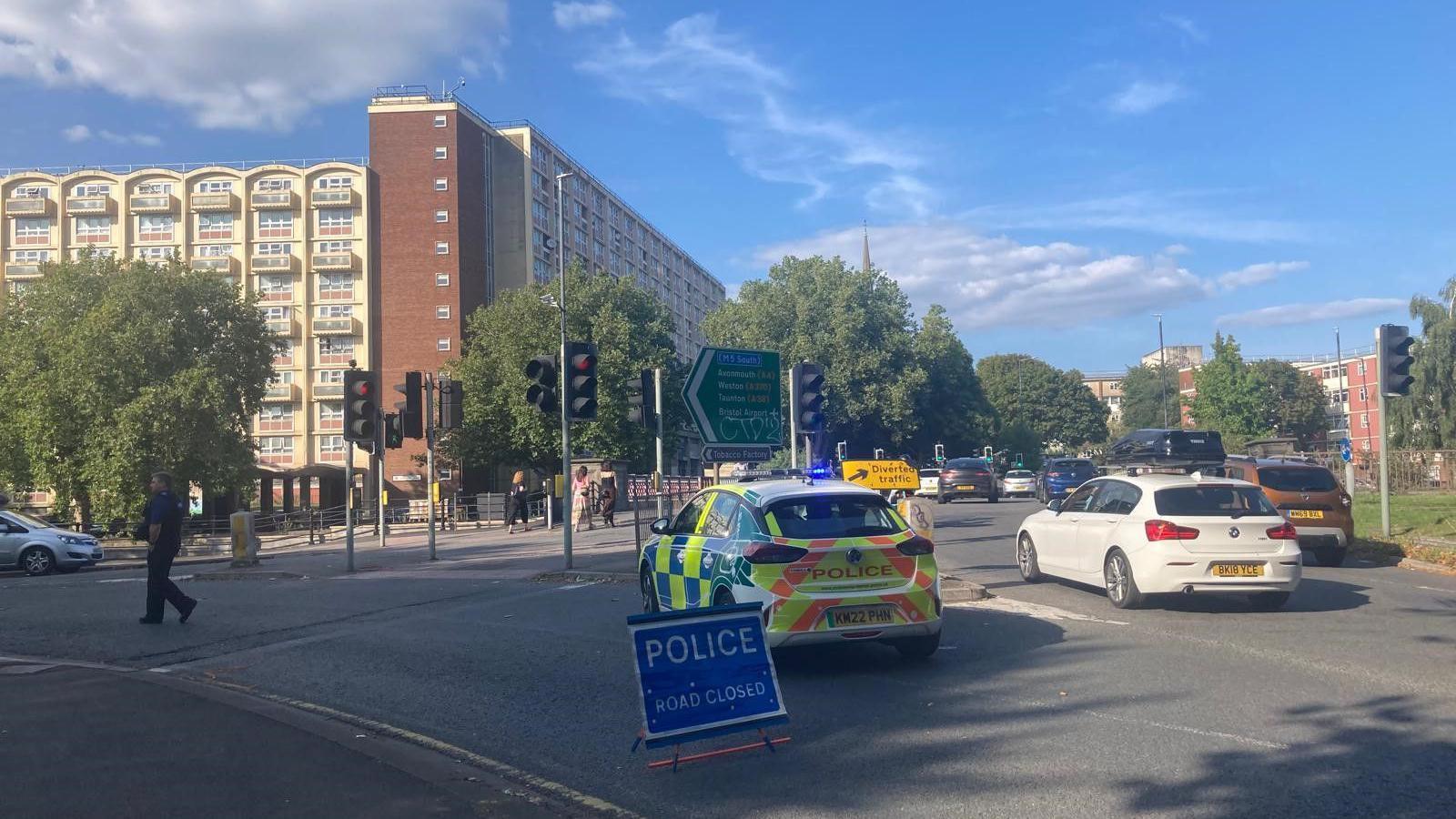 A police car parked on busy Bedminster Bridge roundabout. There is a block of flats visible across the road and lots of cars to the right. The police car is blocking the road and a police officer can be seen walking in front of it. 