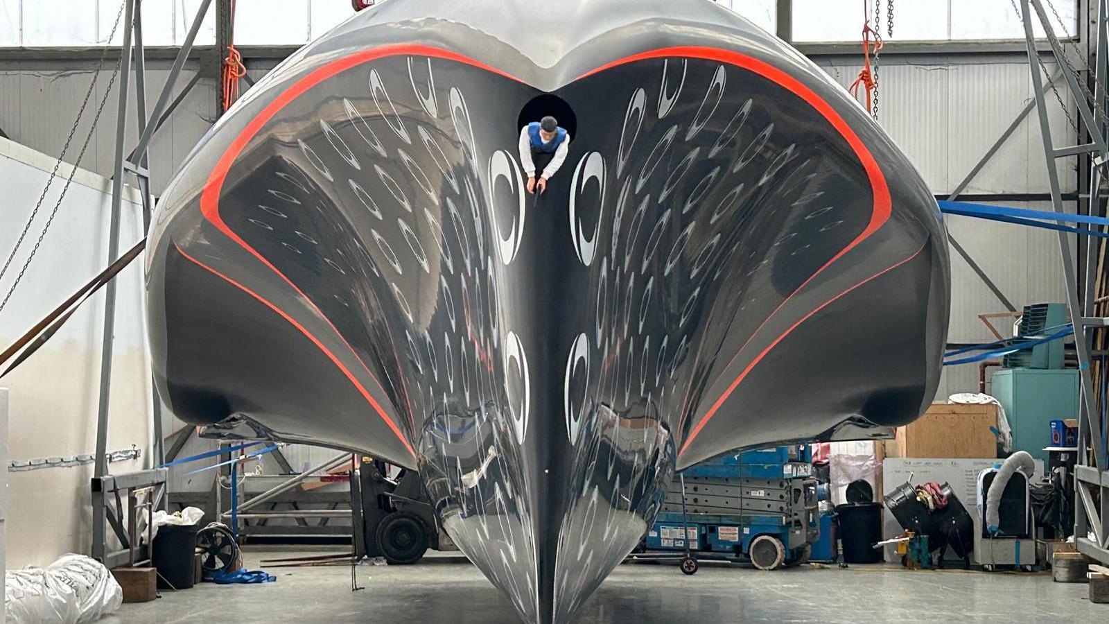 A finished boat hull sat inside the boat at Carrington Boats warehouse in Hythe. It's filling the frame and is blue with silver INEOS logos dotted all over it. There is a model-figurine sticking out of a hole in the bow of the boat