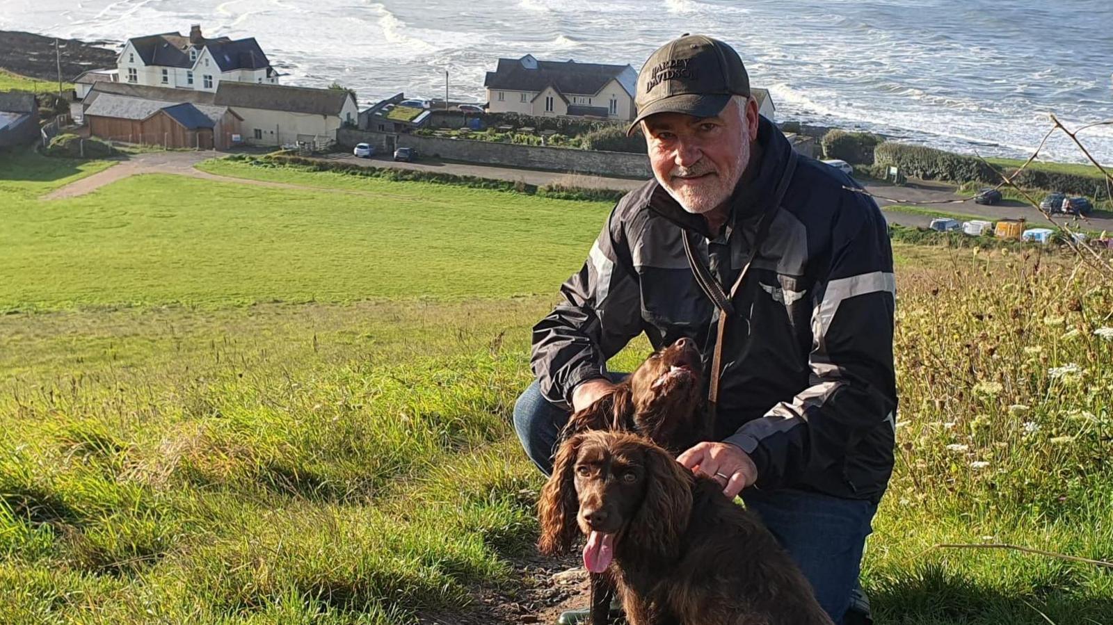 Keith Steer kneeling down beside two brown spaniels on a grassy hillside overlooking the sea. He is wearing blue jeans, a black raincoat and a black cap.