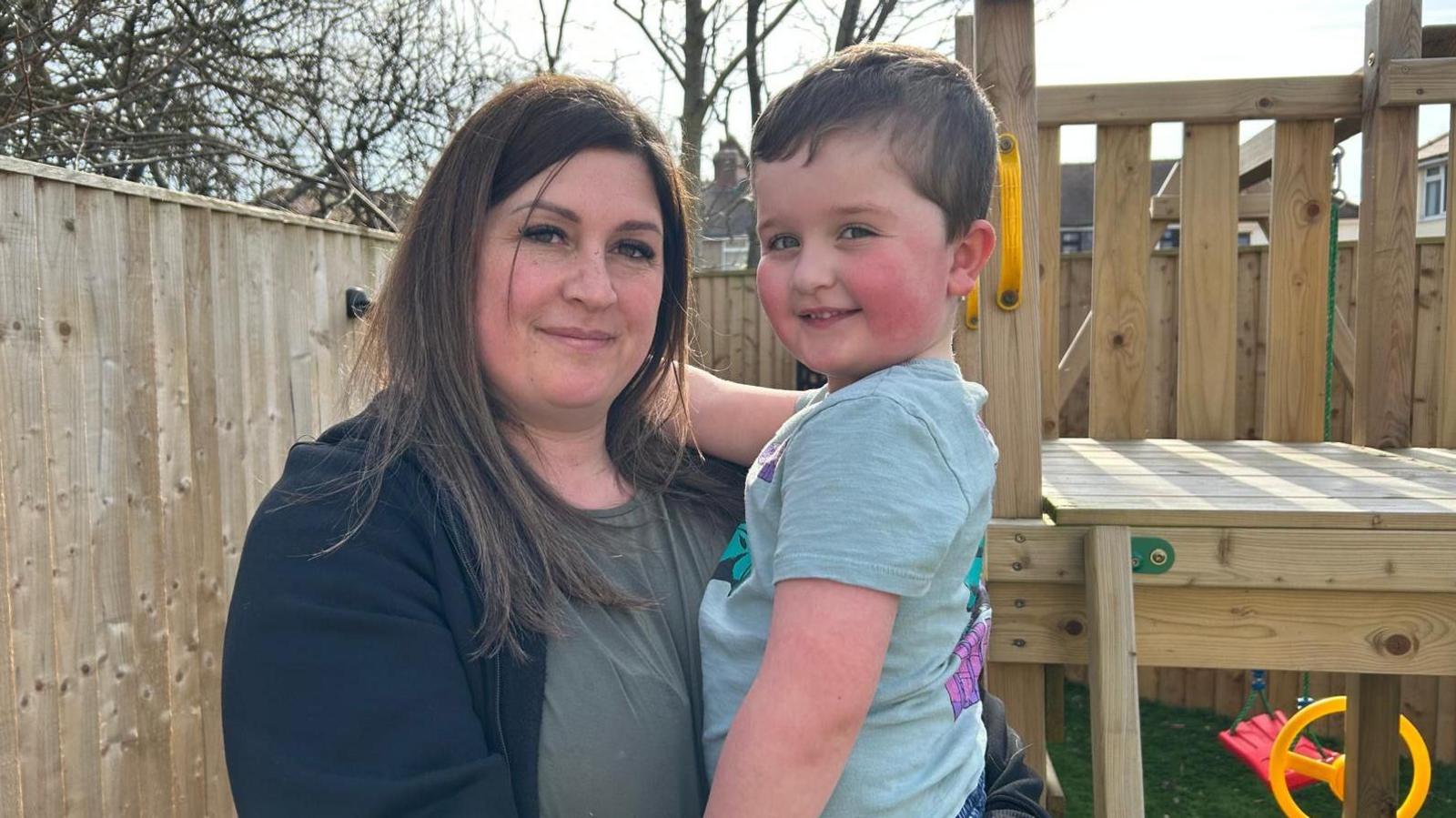 A woman with brown long hair, holding a four-year-old boy, in a garden. A wooden playground and wooden fence is behind them. It is a sunny day.