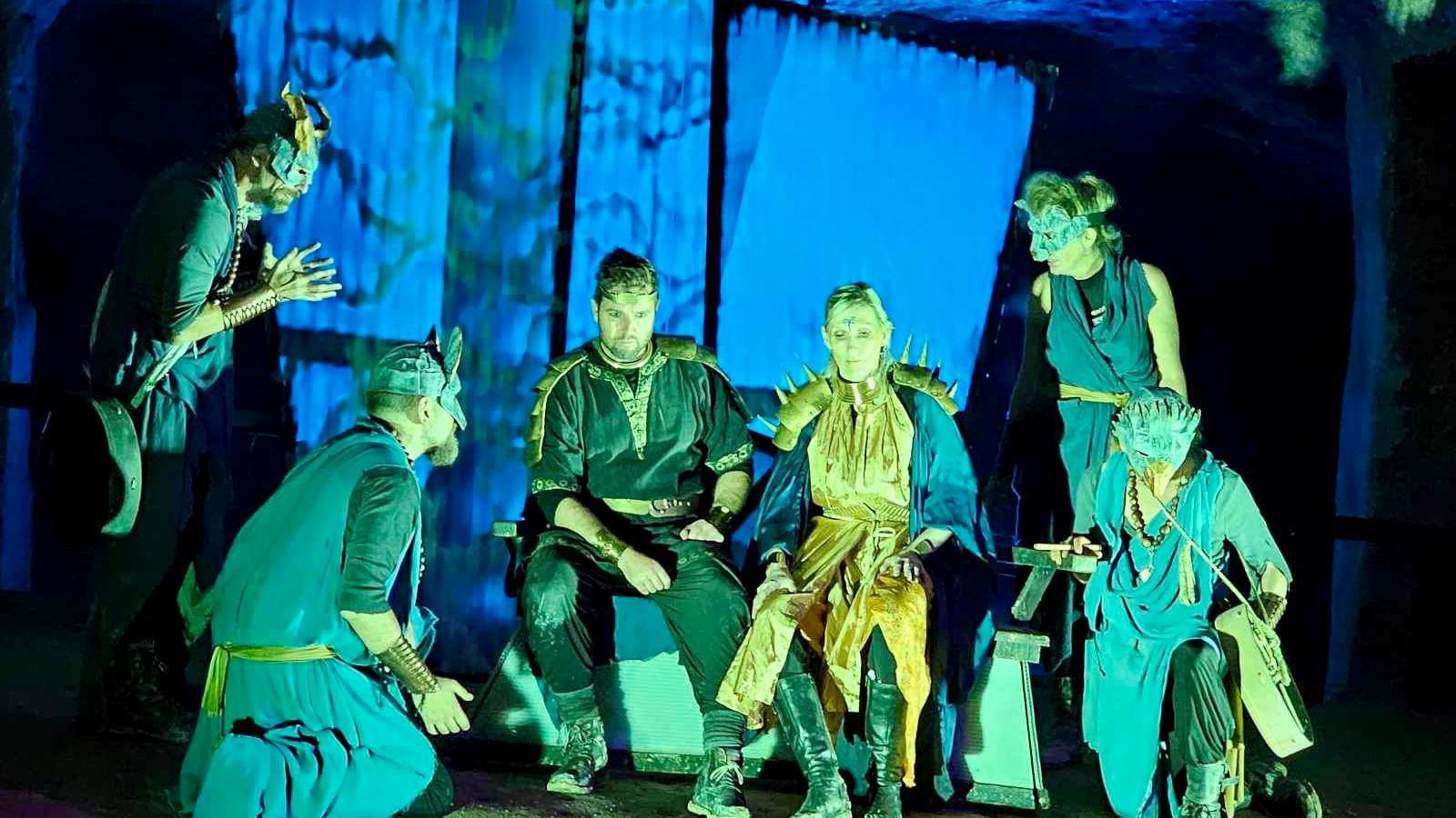 Six people on stage which is lit with a blue-ish light, each person is wearing a costume, some with masks on.