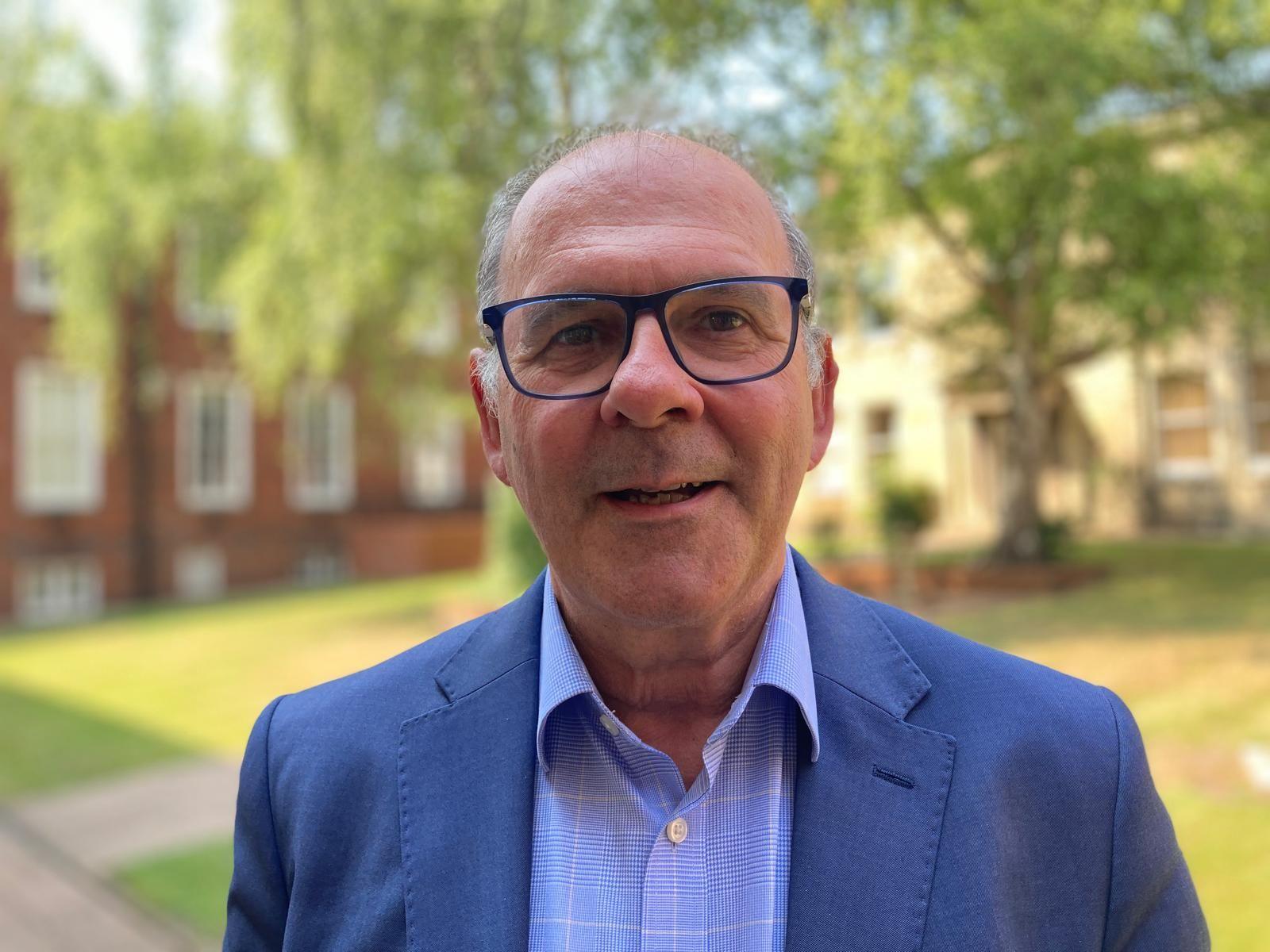 Prof Neal Juster is wearing a blue suit and a lighter blue shirt, he has dark framed glasses and grey hair. He is standing in a lawned courtyard with a tree in the centre.