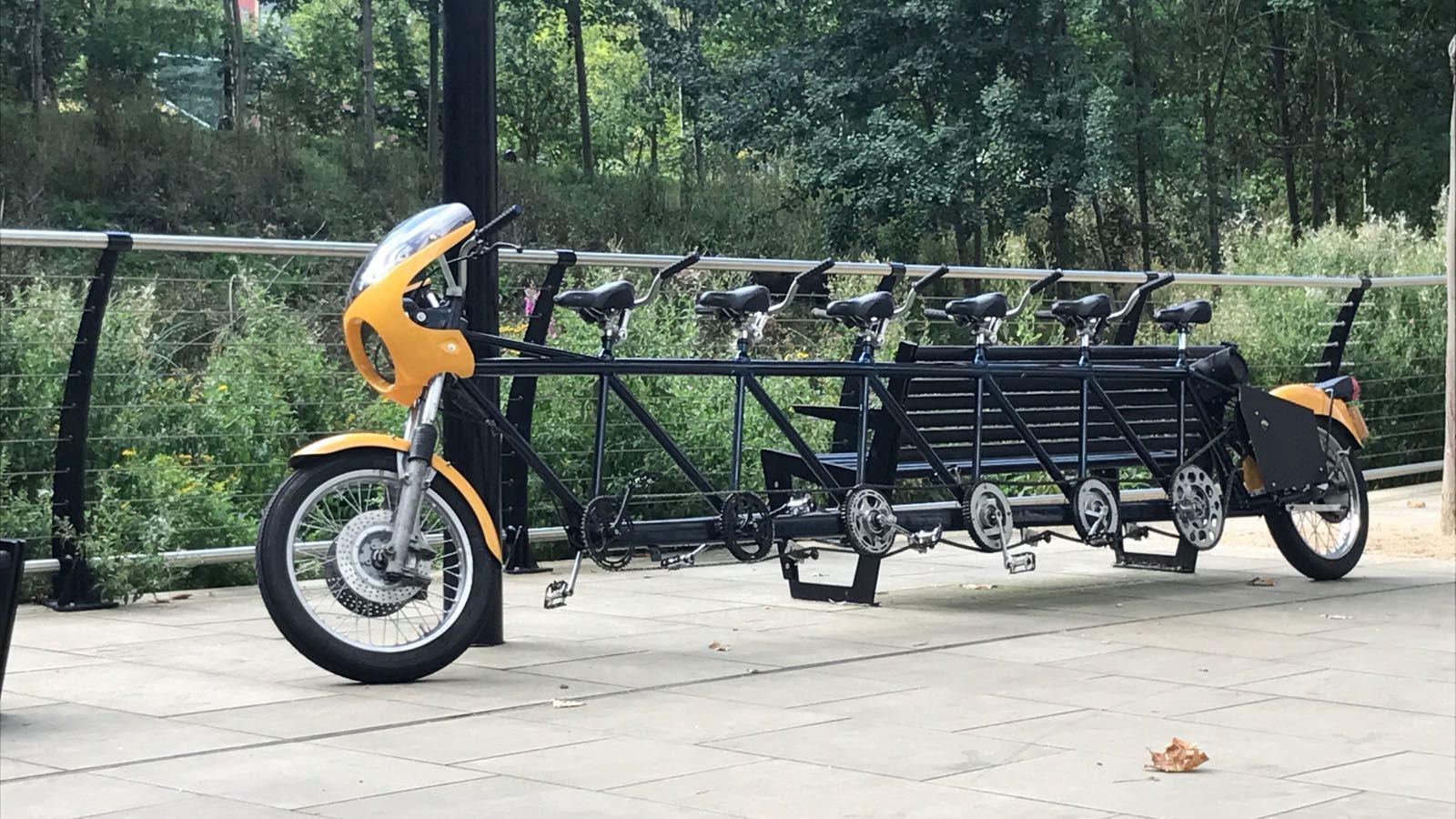 A very long bicycle capable of carrying six people resting against a park bench. There is a metal fence and trees and bushes in the background.