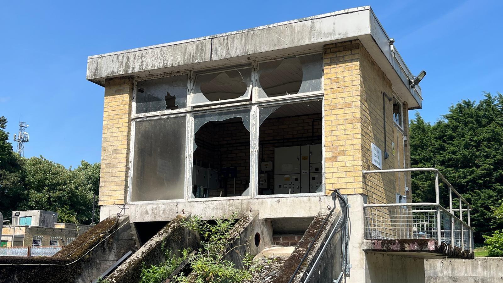 A small building housing machinery with yellow brick walls and three large windows, two of which have been smashed
