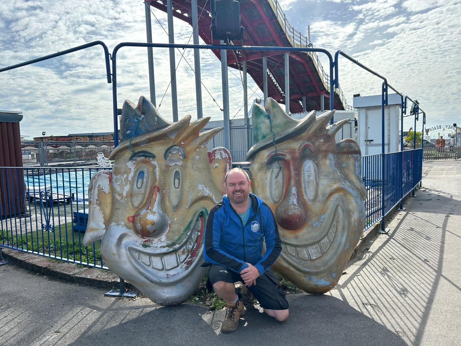 A man in a blue top crouching down between two large vintage clown heads with peeling paint on, in front of a fairground ride